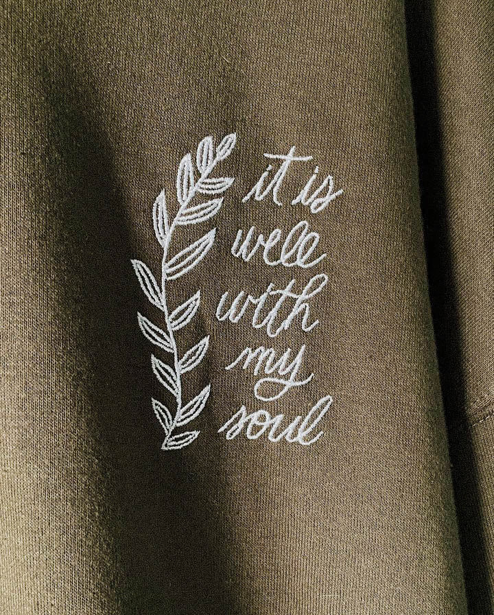 Close up of It Is Well With My Soul embroidered on a dark green hoodie.